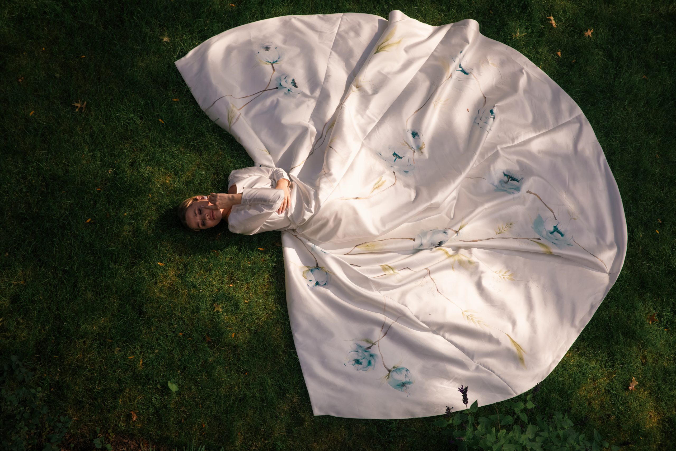 model laying on green grass with silk mikado bridal dress with blue peony hand painting